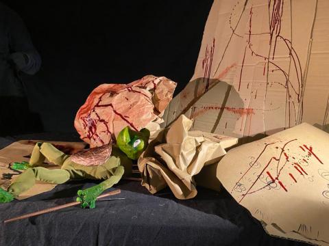 A black stage with cardboard painted red to look like blood, and a costume of a green frog lying supine