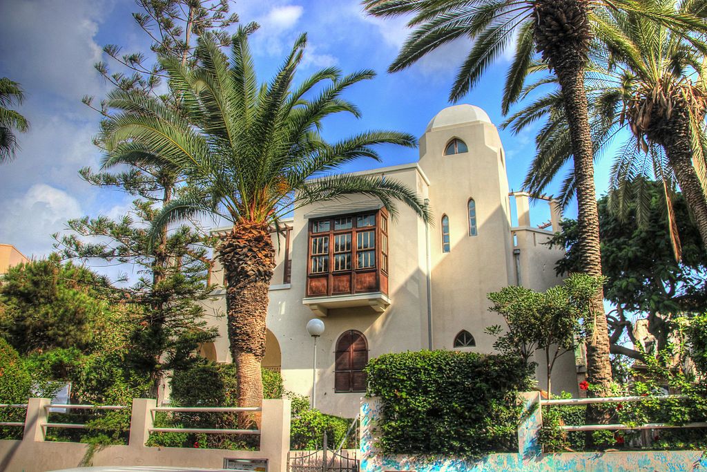 A tall, beige building with brown wooden windows and door is surrounded by palm trees and greenery. The sky is bright blue with clouds.