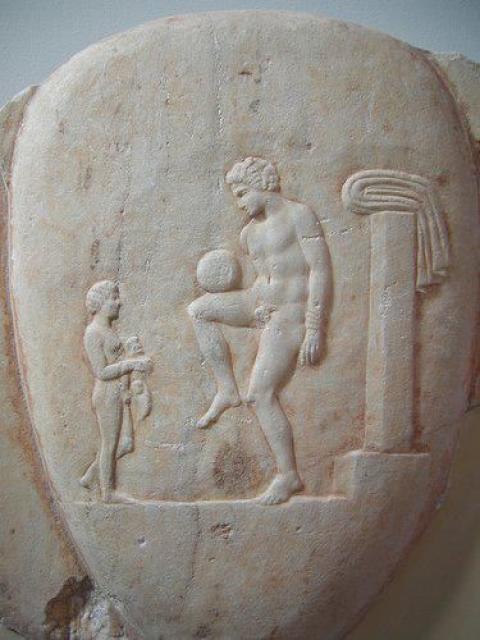 Ancient Greek football player balancing the ball. Part of a marble grave stele, found in Piraeus, 400-375 BC. Item (NAMA) 873 of the National Archaeological Museum, Athens. Image via Wikimedia under Public Domain.