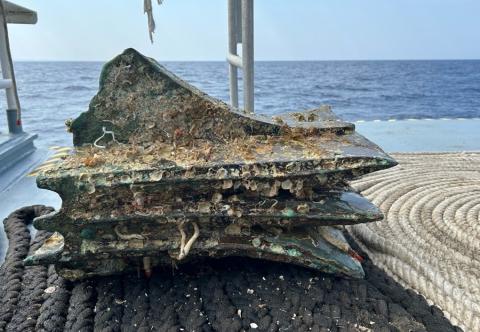 A large metal object covered in barnacles and seashells sits on top of a bed of ropes aboard a ship