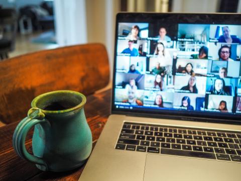 A Macbook sits on a wooden desk showing a Zoom screen filled with faces. Left of it, a turquoise mug sits on the desk.
