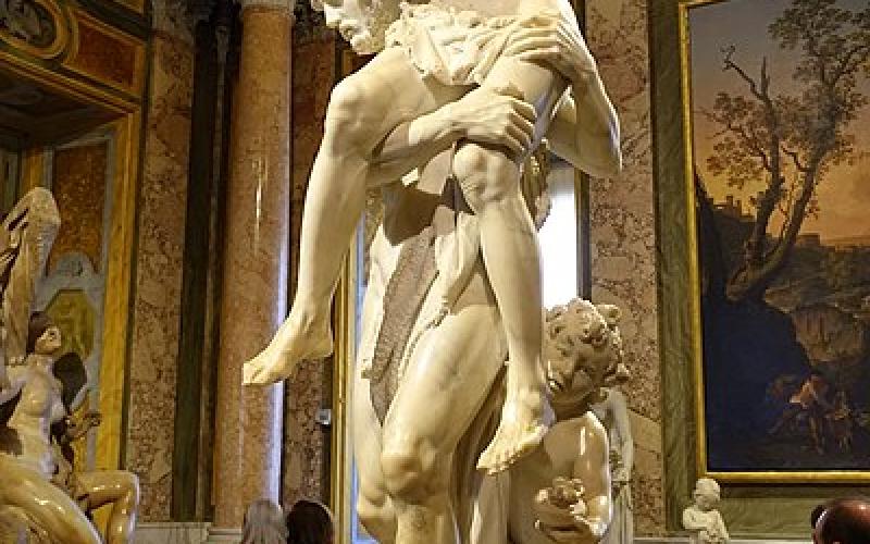 A white marble statue of a nude man holding a smaller old man on his shoulder with a child behind his legs. The old man carries a statue.