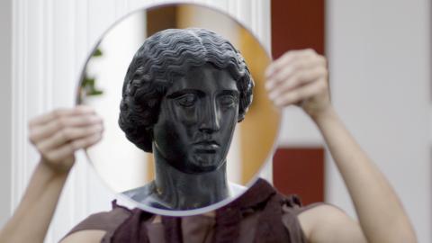 A woman holds a round mirror in front of her head. In the mirror, we see a black sculptural head of an ancient woman.