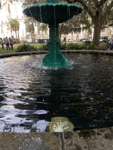 A green fountain surrounded by a basin of water. In the basin is a gold piece with a woman's left hand on it, the fingers apparently severed.