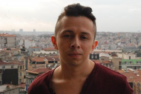 A man with short hair and a maroon t-shirt faces the camera with a city in the background