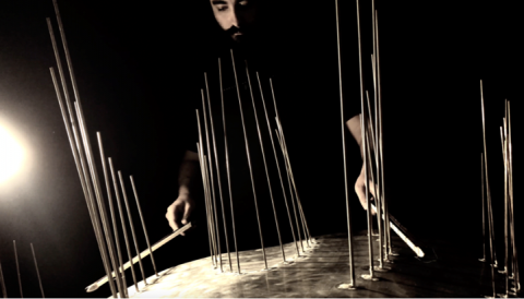A dark image of a bearded man standing over a metal plate, from which a number of vertical rods are extending. He is hitting the rods with two drumsticks.