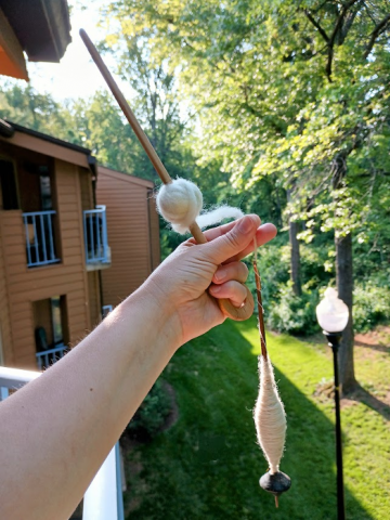A person's left hand holding a wooden rod with a ball of white yarn attached to it. Between the fingers is a small wooden spindle with yarn wrapped around it.