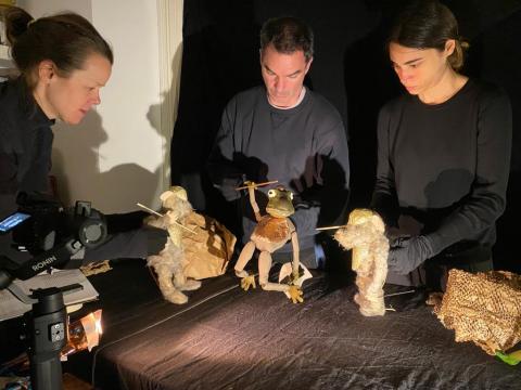 Three people dressed in all black hold puppets of mice and frogs against a black backround while a camera films.