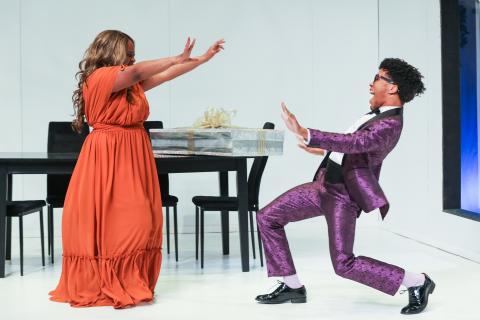 A black woman in an orange dresses raises both her hands at a black man in glasses and a purple suit, who grins back at her while bending his knees
