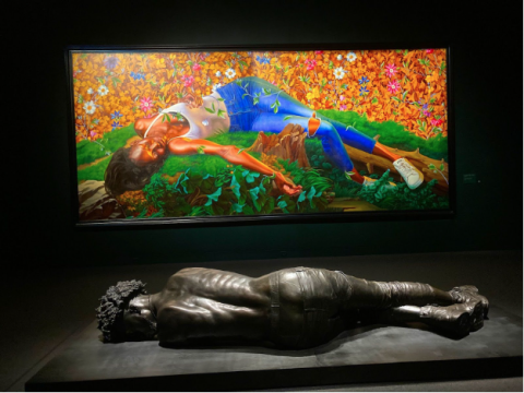 A horizontal bronze statue of a shirtless person with curly hair, jeans, and sneakers lies in front of a brightly-painted canvas depicting a black person lying horizontally on a grassy hill wearing a white tank top, blue jeans, and sneakers.