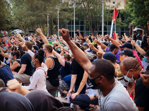 A crowd of people facing left with their right arms raised making fists. Many of them wear face masks. There are trees in the background.