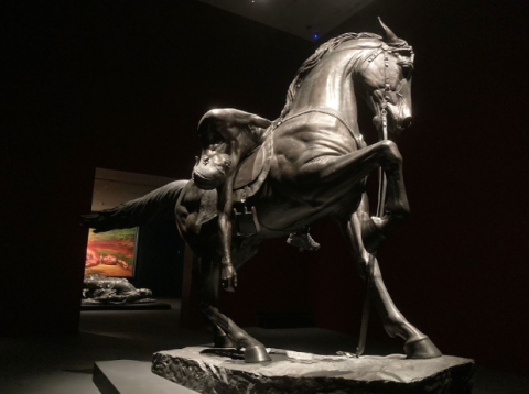 A dark room with light shining onto a bronze statue of a horse with its right leg raised. Across the horse's back, a shirtless black man lies with eyes closed and one arm hanging towards the ground.