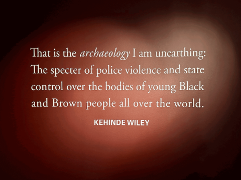 A black wall with light shining on text that reads "That is the archaeology I am unearthing: The specter of police violence and state control over the bodies of young Black and Brown people all over the world. Kehinde Wiley"