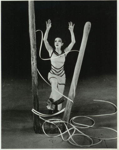 Black and white photo of a woman in a dress with her arms raised standing between two wooden poles with rope wrapped around them
