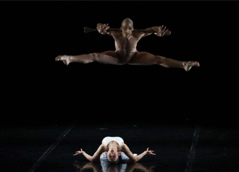 A nude male figure jumps in the air with arms and legs outstretched. Beneath him, a woman in white lies on the floor bent over backwards.