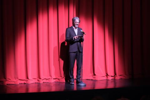 A man with gray hair and glasses wearing a dark suit stands on a stage in front of a microphone reading off note cards. A red curtain is behind him and a spotlight shines on him.