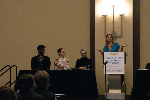 A woman in a blue blouse stands at a podium. Next to her, three panelists sit behind a long table.