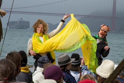 A woman wearing and holding a long yellow scarf raises on arm next to a woman in a pig mask. With the Golden Gate Bridge behind them, they face an audience, all on a boat.