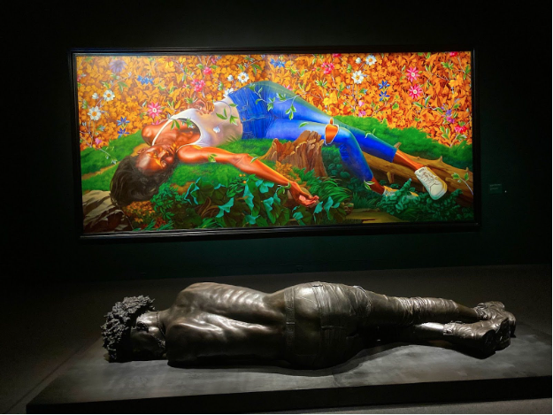 A bronze statue of a shirtless black person wearing jeans lies horizontally, facing away from the camera. Behind it is a brightly-colored canvas with a painting of a young black woman lying on her back, eyes closed, arms spread, in a white tank top, jeans, and sneakers. Behind the person, the background is covered in yellow and red flowers.