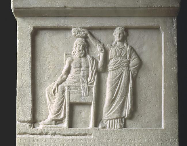 A marble relief depicting man wearing a toga sitting in a chair while a woman stands behind him placing a crown on his head.