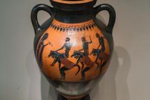 A black-figure vase depicting three chorus-men costumed as warriors, wearing individually crested helmets, “riding" three partners in horse costume.