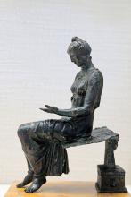 A bronze statue of a girl sitting on the side of a bench in reading pose, though she does not hold a book. Her hand is open as if a book is missing. She is barefoot, her hair tied up, wearing a draped dress.