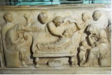Stone relief in which the body of a child lies on a couch, surrounded by people in various gestures of mourning.