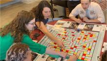 People sit around a table playing a board game. Two women on the left reach their arms across the board. One is pointing with her index finger.