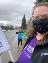 A white woman wearing rectangular glasses, a black mask, and a purple t-shirt holds a white flag. Behind her, a person in a black jacket with a fur-trimmed hood holds a sign. They are outdoors on the sidewalk, and the sky is cloudy.