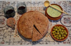 A dinner spread atop a mosaic-printed tablecloth. Two small glasses of red wine, a round bread loaf sliced into eighths, a terracotta bowl of green olives, and a bowl of pesto with a wooden spoon.