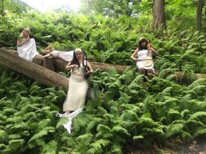 Four women in white tunics sit and lie in a landscape of ferns. Some play instruments.