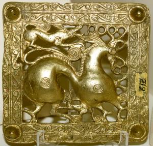 An ornate carved gold square, at the center of which is a stylized horse with a small winged animal resting on its hind quarters. There are decorative patterns forming a border around the horse.