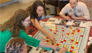 People sit around a table playing a board game. Two women on the left reach their arms across the board. One is pointing with her index finger.