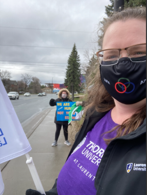 A white woman wearing rectangular glasses, a black mask, and a purple t-shirt holds a white flag. Behind her, a person in a black jacket with a fur-trimmed hood holds a sign. They are outdoors on the sidewalk, and the sky is cloudy.