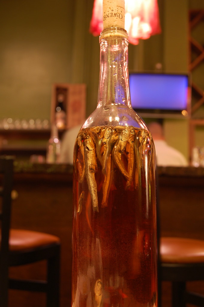 A tall, glass bottle filled with a brownish-red, clear liquid. At the top floats a cluster of sardines.