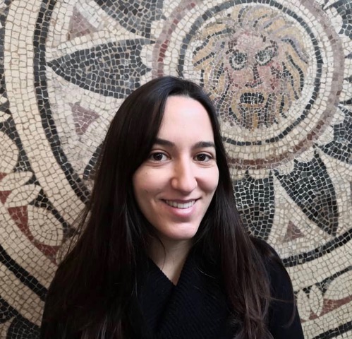 A woman with long, black hair smiling at the camera. She is wearing a black sweater and standing in front of a Roman mosaic that depicts a man's head with light hair and an open mouth.
