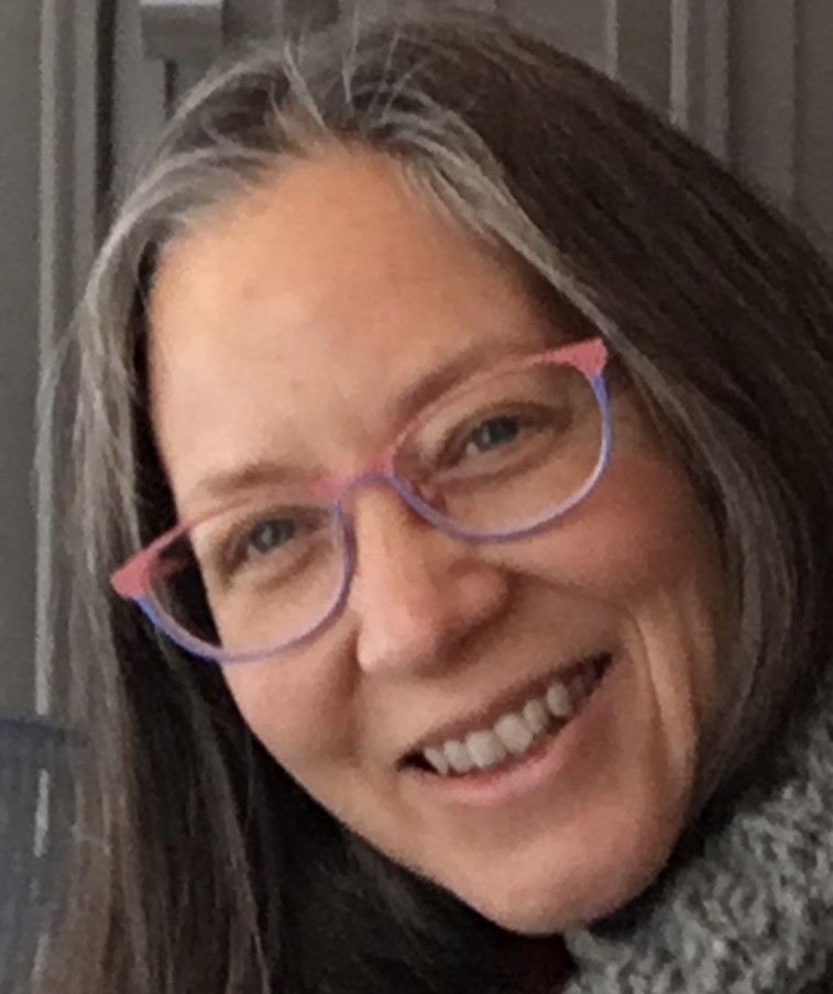 A light-skinned woman with gray hair and pink/purple glasses smiles at the camera.