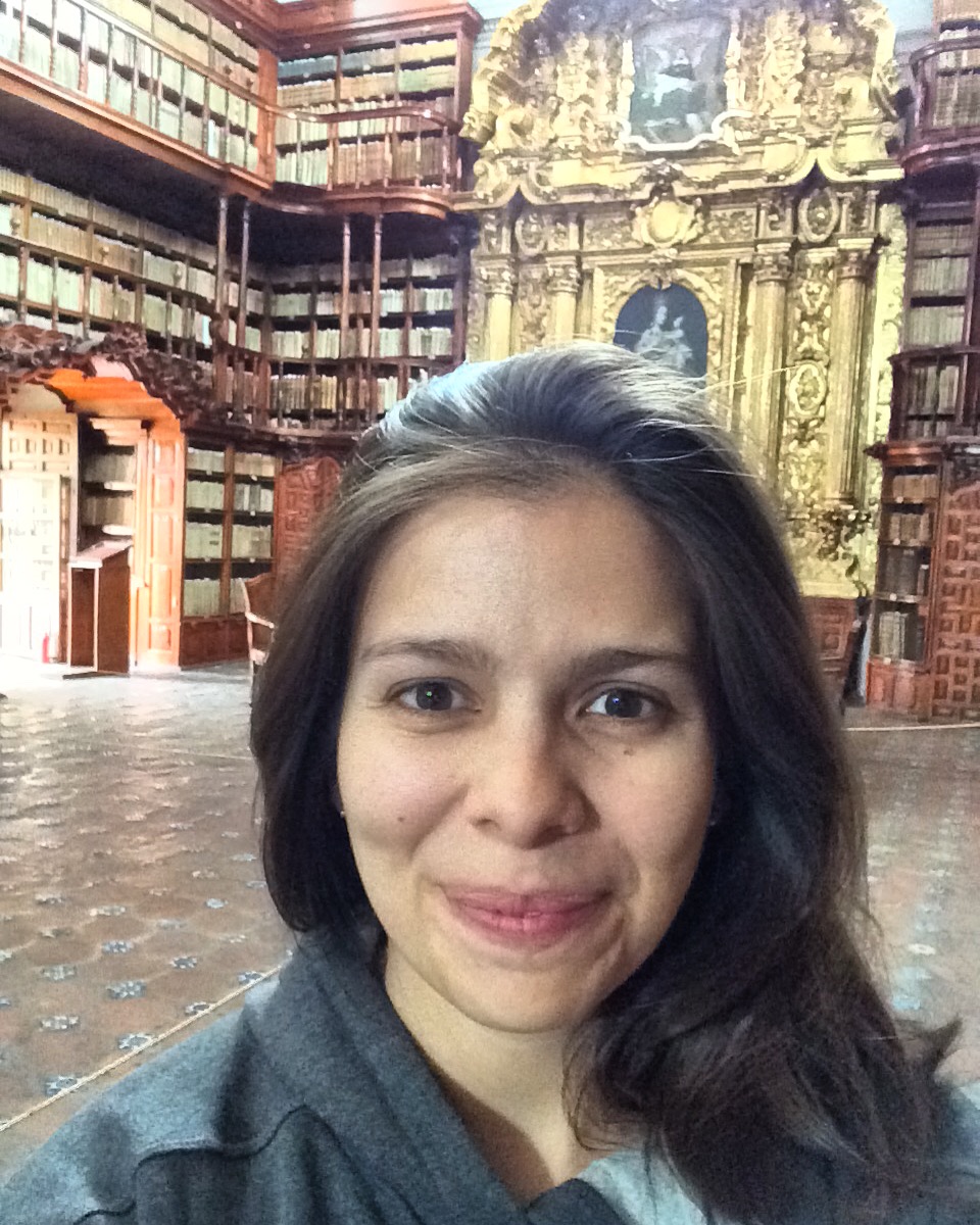 A woman with dark brown hair smiling at the camera. Behind her is a large room lined with tall wooden bookshelves filled with books.