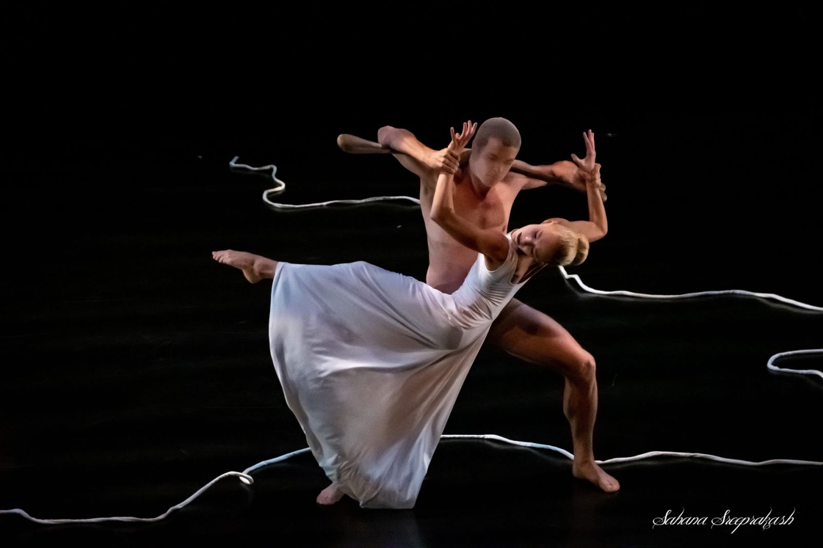 A nude man and a woman in a white dress engaged in modern dance against a black background. A white string wraps around behind them.
