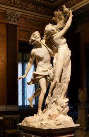 A white marble sculpture of Apollo chasing Daphne. Apollo is wearing a tunic loosely around this waist and reaches his left hand around Daphne's waist. Her cloak is falling off, her right hand is raised above her head, and her long curly hair is turning into tree branches at the ends.