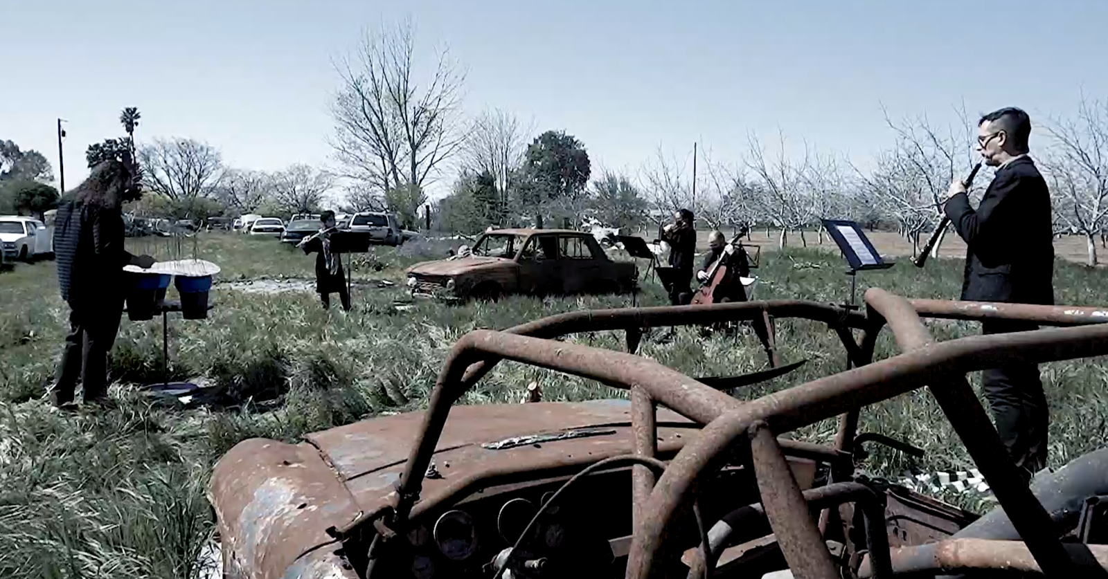 A grassy field with cars parked in the background. Five figures in all black stand apart from each other playing various instruments. The rusted chasses of old cars are strewn throughout the grass.