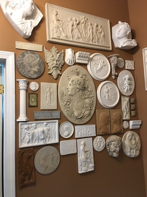A tan wall with a collection of white bas-relief sculptures hanging on it. Most depict wither a bust or a face, or an entire person or mythological creature. There are also some columns and decorate elements.