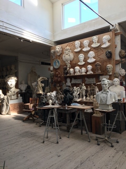 An art studio with a wooden floor and a number of small tables, upon which sit small plaster statues and busts. Sculpted heads are lined up on a shelf, with some hanging on a wall behind it.