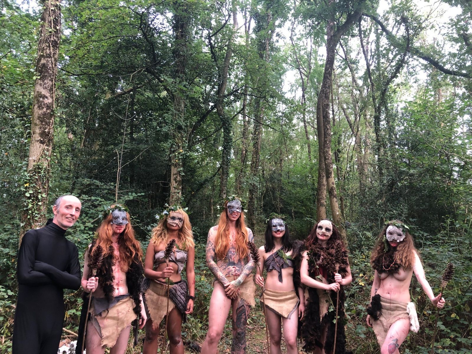 Six women stand in a forest in various costumes and masks of leaves and natural fabrics covering portions of their bodies. On the left stands a man in an all-black bodysuit.