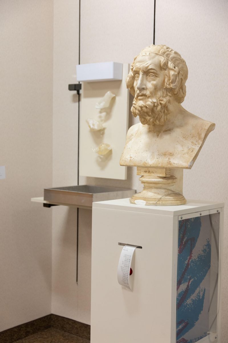 A white marble bust of a man with shaggy, curly hair sits atop a white pedestal. A receipt is coming out of an opening on the pedestal, and the side of the pedestal is decorated with a teal and purple zigzag pattern that used to appear often on paper cups in the 90s.