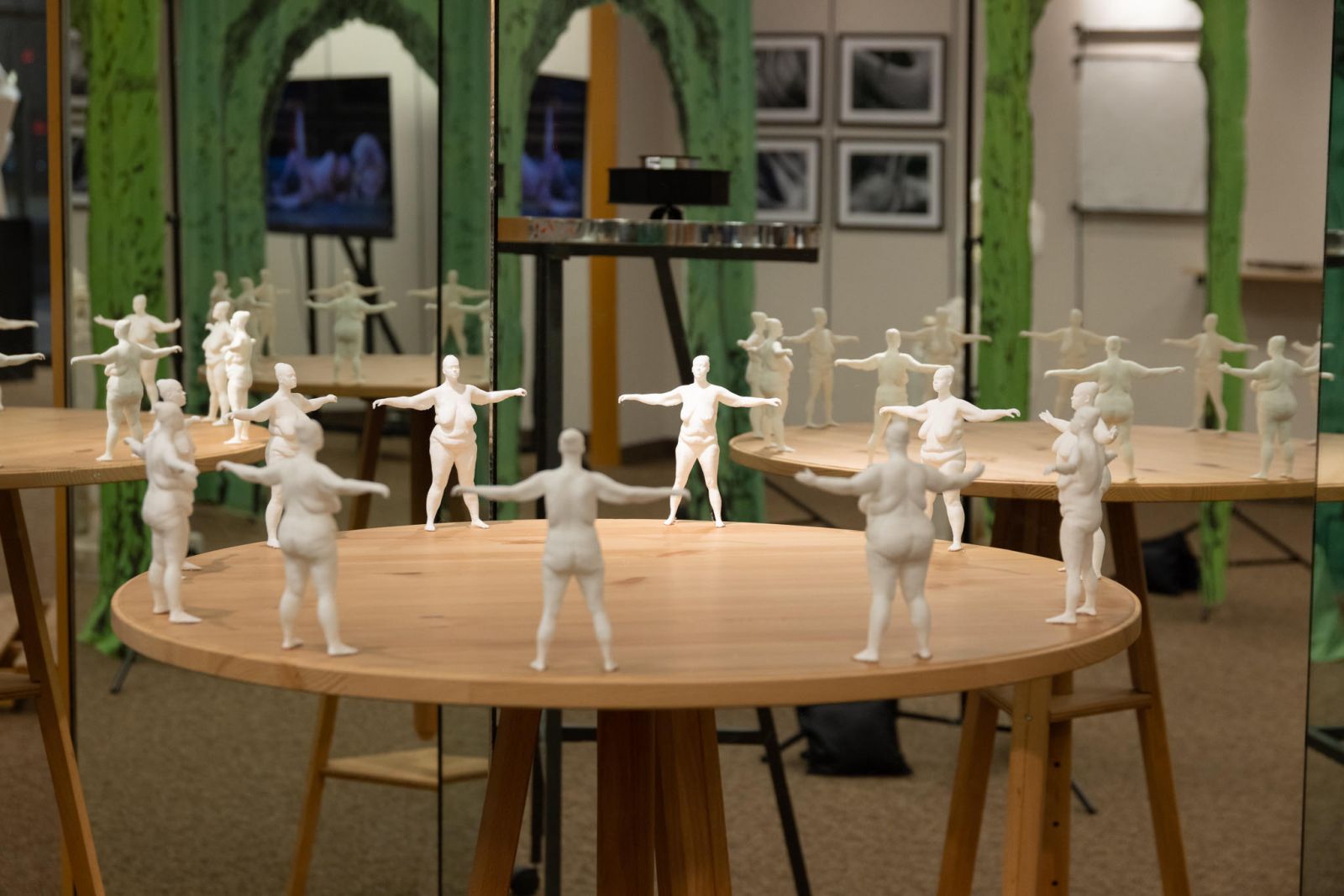 A series of round, wooden tables atop which stand small, white sculptures of nude women with their arms extended to the sides.