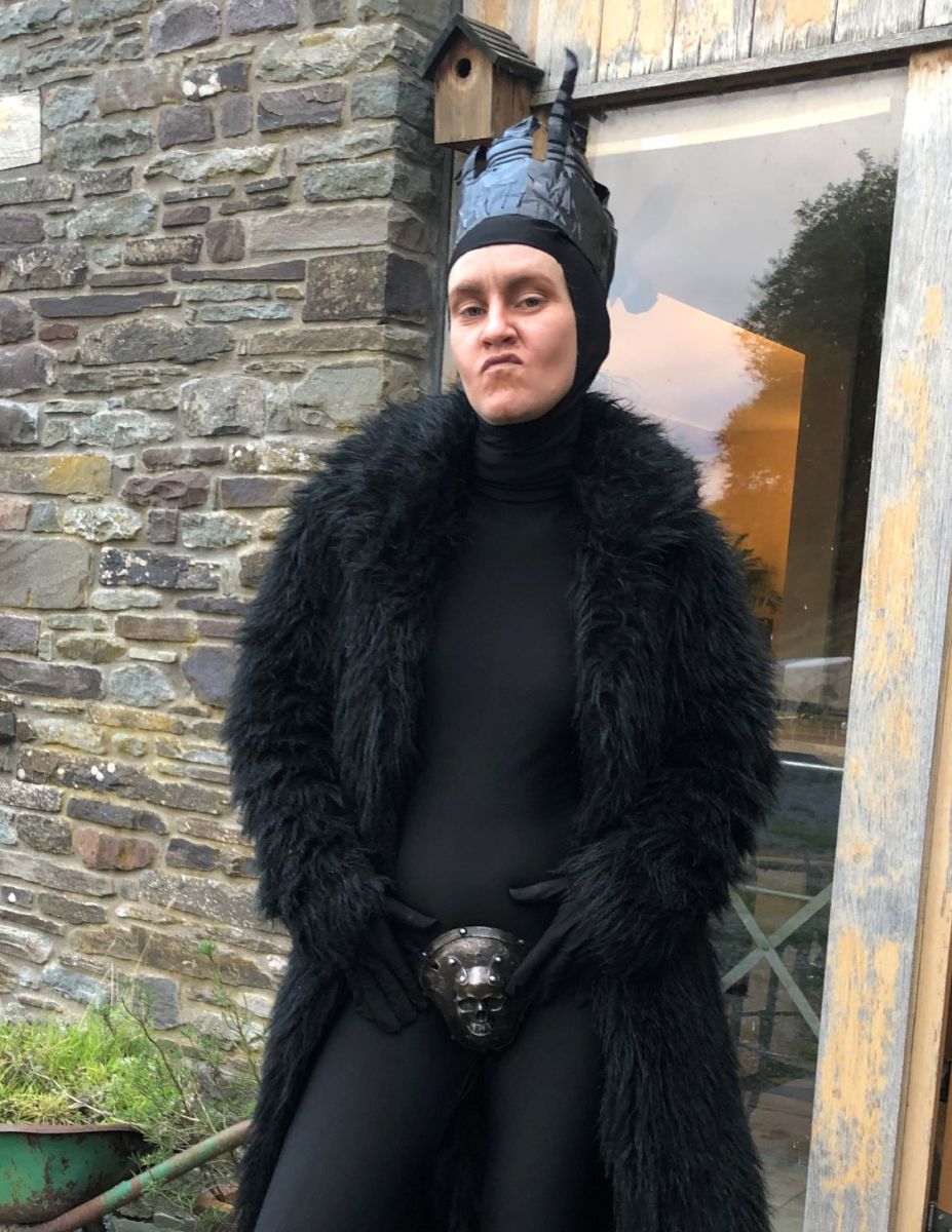 A woman stands in front of a brick building wearing an all-black body suit with a black faux fur coat on top, black gloves, a gray crown-like hat, and a silver skull over her crotch. Her hair is hidden in a hood, and she is sneering at the camera.