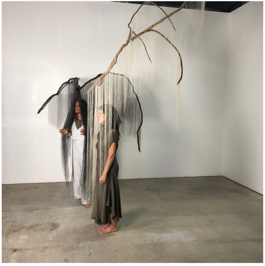Two women, one a long white dress and one with gray, stand in an empty, white-walled studio with large branches dangling over their heads. From the branches hang many thin threads that cover the women's heads.