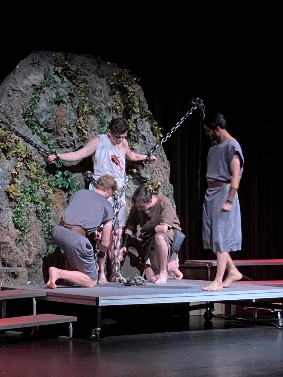 A man in a white tunic with a red blood stain over the chest stands in front of a moss-covered rock, his arms straight out, his wrists shackled to chains. Two men kneel on either side of him binding his feet. A third man stands watching them.