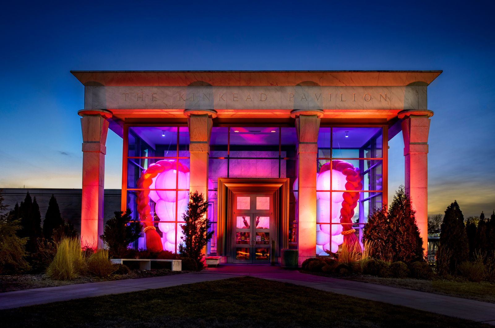 A stone building with greek-style columns is lit up at dusk. Inside it, you can see bright, lit-up sculptures: two red braids and two bunches of pale pink spheres.